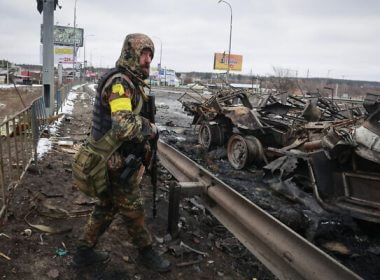 An armed Ukranian man stands by the remains of a Russian military vehicle in Bucha, close to the capital Kyiv, Ukraine, March 1, 2022. (AP Photo/Serhii Nuzhnenko)