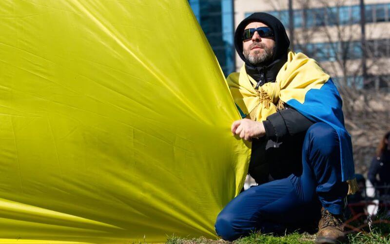 A man wearing sunglasses, a hoodie and a Ukrainian flag around his shoulders squats during a rally in Farragut Square, Washington, D.C., Feb. 27. (Colin Demarest/C4ISRNET)