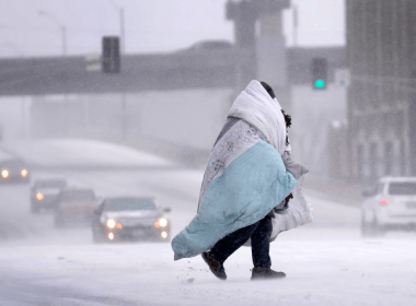 A person wrapped in a blanket crosses a snow-covered street, Dec. 22, 2022, in St. Louis. Snow and frigid temperatures have moved across the U.S., complicating holiday travel and plans in the coming days.