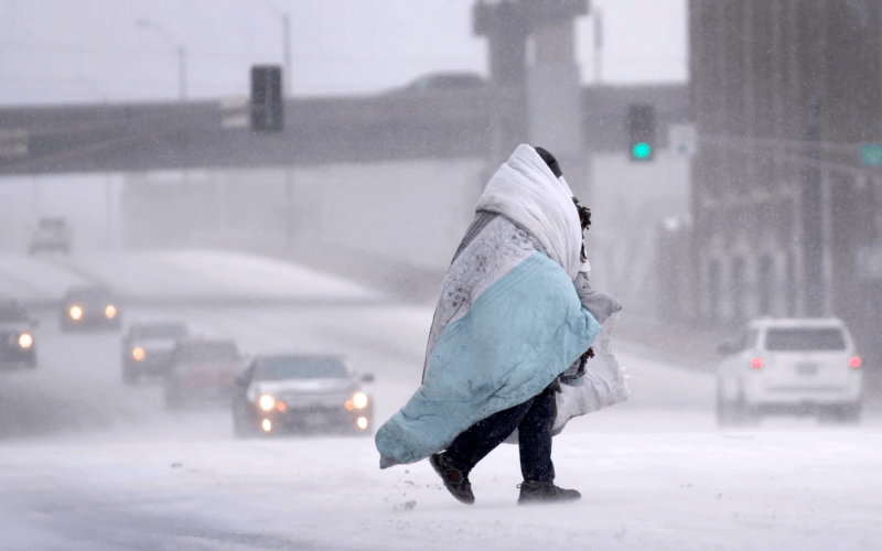 A person wrapped in a blanket crosses a snow-covered street, Dec. 22, 2022, in St. Louis. Snow and frigid temperatures have moved across the U.S., complicating holiday travel and plans in the coming days.