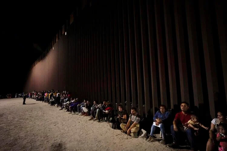 Migrants wait along a border wall Aug. 23, 2022, after crossing from Mexico near Yuma, Ariz. U.S. immigration offices have become so overwhelmed with processing migrants for court that some some asylum-seekers who crossed the border at Mexico may be waiting a decade before they even get a date to see a judge. Gregory Bull / AP Photo